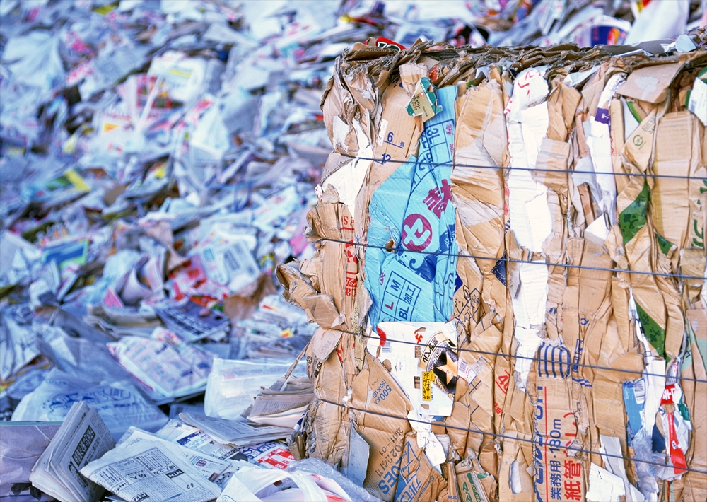 IMAGE_この写真は、リサイクルされた紙や段ボールが積まれている場所を示しています。全体的に多くの紙くずや梱包材が見えており、圧縮された段ボールの束も含まれています。これは、おそらく廃棄物処理やリサイクルのための一時保管エリアかもしれません。