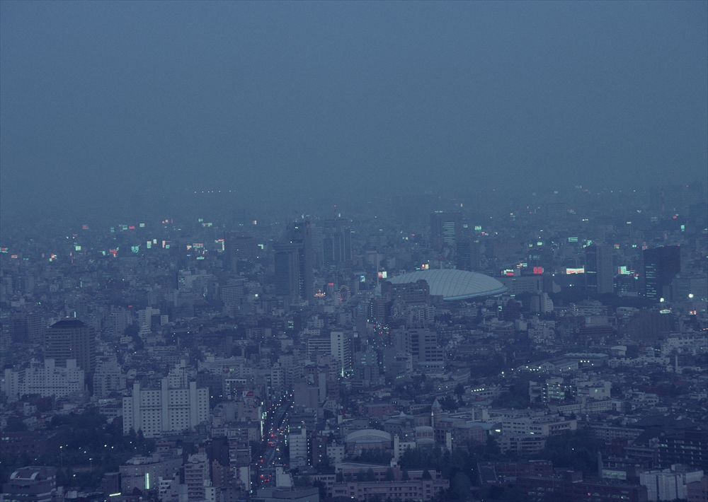 IMAGE_これは都市の夜景や夕暮れ時の風景の写真のようです。多くの建物や明かりがあり、霧や雲に包まれているために少しぼんやりとした印象を与えています。都市の全体的な雰囲気や建物の形状から、大都市である可能性があります。