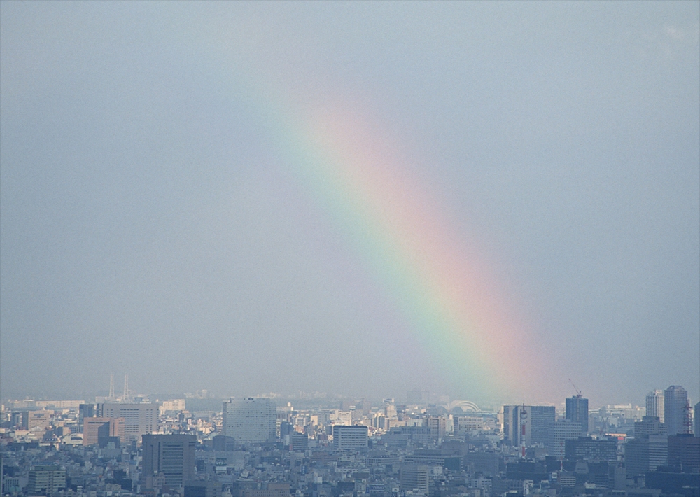 IMAGE_これは、都市の風景に虹がかかっている写真です。空には薄い雲があり、虹はその上に現れています。このような風景は自然の美しさを感じさせ、晴れた後の特別な光景として人気があります。