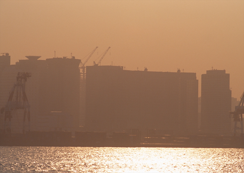 IMAGE_これは、夕暮れ時の港の風景を捉えた写真のようです。シルエット状の建物や工事用クレーンが見え、反射した水面が印象的です。全体的に柔らかい色合いで、穏やかな雰囲気がありますね。
