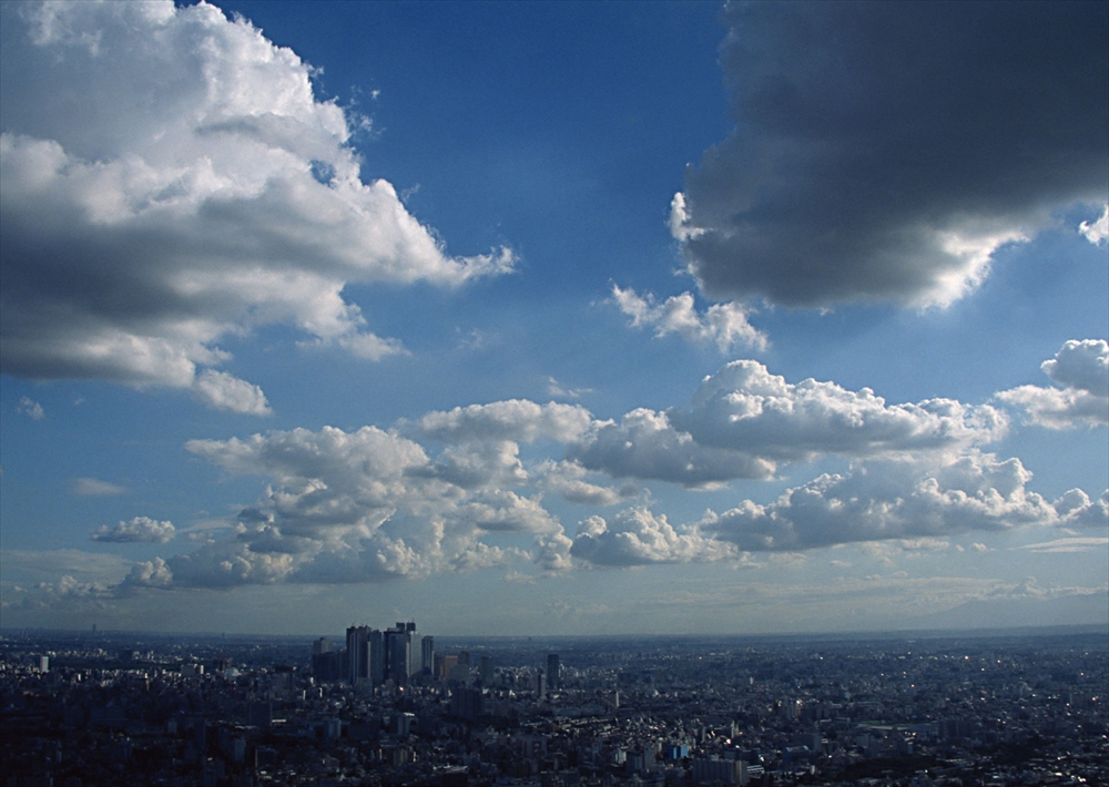 IMAGE_これは都市の風景と空を捉えた写真のようです。高層ビルが見え、その奥には広がる雲と青空があります。このような風景は、都市の上空や高い場所からの眺めを示していると思われます。