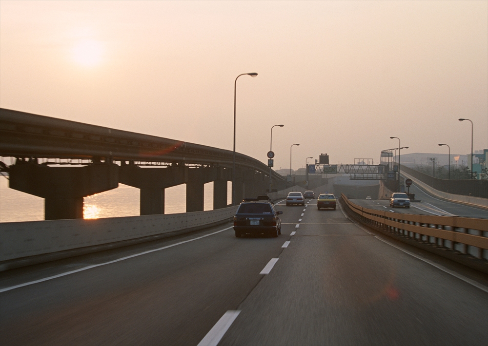 IMAGE_この写真は、高速道路や道路の風景を捉えたもののようです。夕暮れ時で、太陽が沈む様子が見え、道路の脇には車や街灯があります。一般的には、都市部の交通の一部を表現した景色と言えるでしょう。