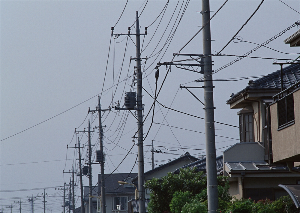 IMAGE_この写真は、電柱とその周辺の住宅が映っている風景です。電線が空に張り巡らされており、建物の一部も見えます。日本の住宅街や町の一角を示しているようですね。