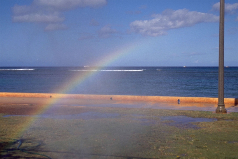 IMAGE_この写真は、海と虹の風景を捉えたものです。青い海と空、そして地面には水が残っている様子があります。虹は海の上にかかっており、自然の美しい景色を感じさせます。横には街灯も見えます。
