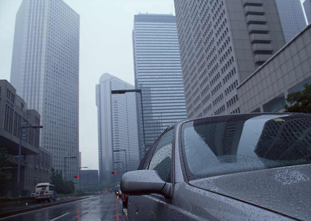 IMAGE_この写真は、雨の中にある都市の風景を捉えているようです。道路には車があり、高層ビルが周囲に立ち並んでいます。霧がかかったような曇り空の下で、都市の風景が少し幻想的な雰囲気を出しています。