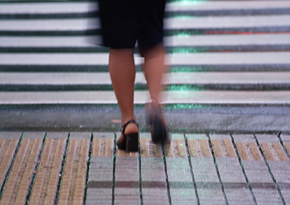 IMAGE_この写真は、交差点を歩いている人の足元を捉えたもののようです。背景には横断歩道のストライプが見え、雨に濡れたような質感があります。全体的に動きが感じられる構図です。