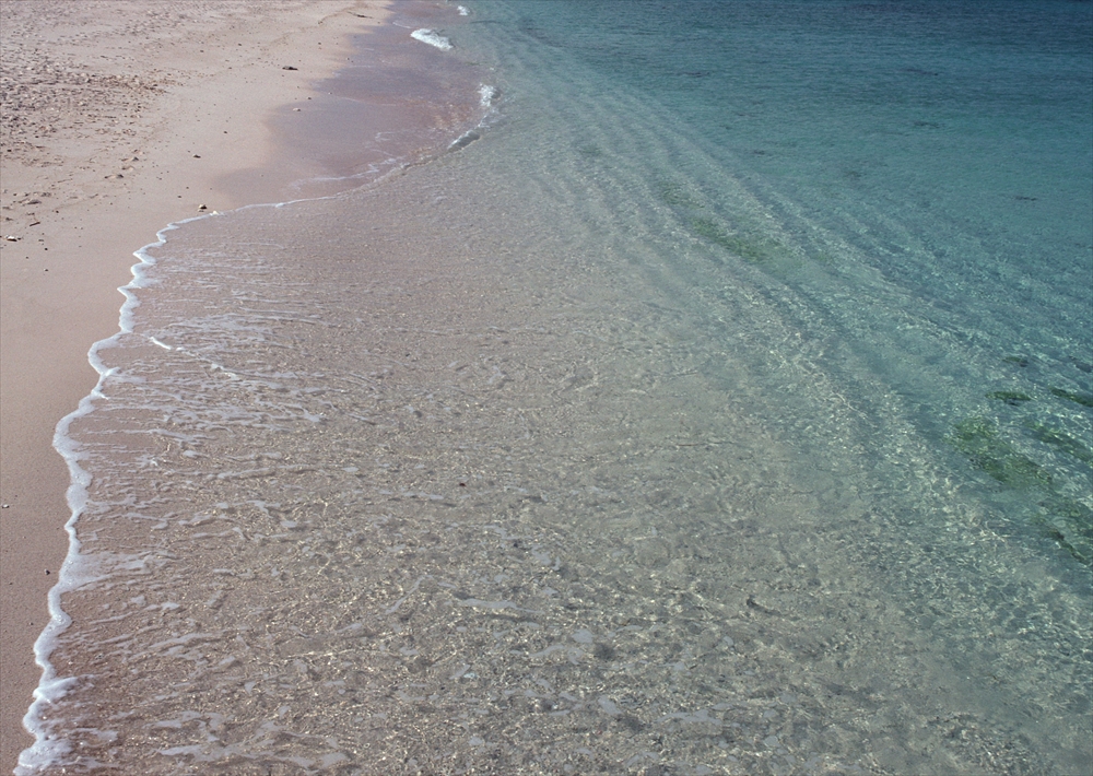 IMAGE_これは砂浜と透明な海の写真です。穏やかな波が海岸に打ち寄せている様子が見えます。全体的にリラックスした雰囲気の風景です。