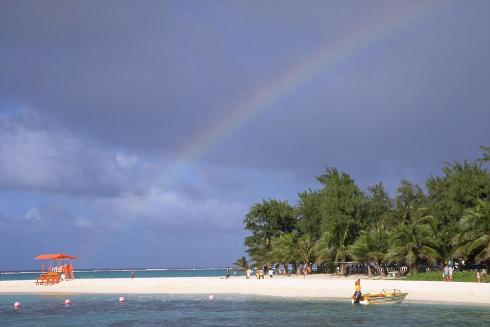 IMAGE_この写真は、ビーチの風景を示しています。虹が空にかかっており、周囲には緑豊かな木々や人々が見えます。また、ボートも海に浮かんでいます。全体として、リラックスできる自然の景色を感じさせるような情景です。