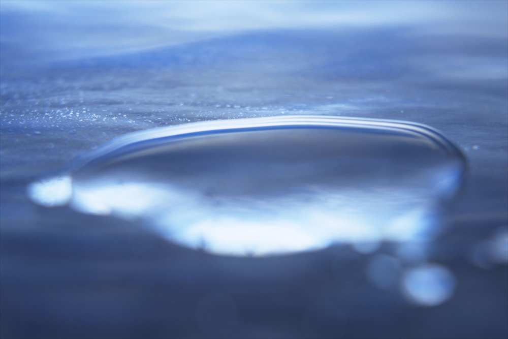 IMAGE_これは、水滴のクローズアップ写真のようです。水の表面を捉えたもので、色合いは青みがかったトーンになっています。水滴の形とその周囲の光の反射が印象的です。